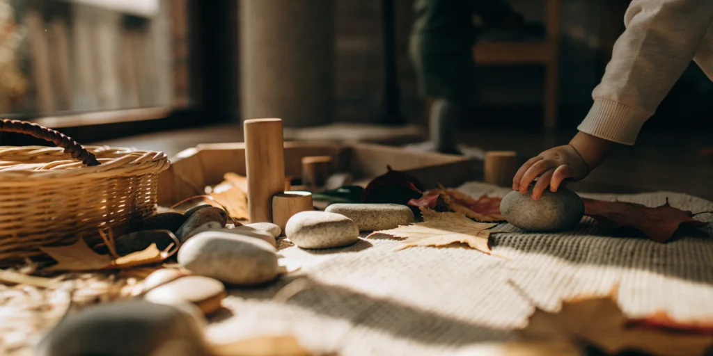 Child’s hand exploring a natural Reggio Emilia provocation with stones, leaves, and wooden blocks.